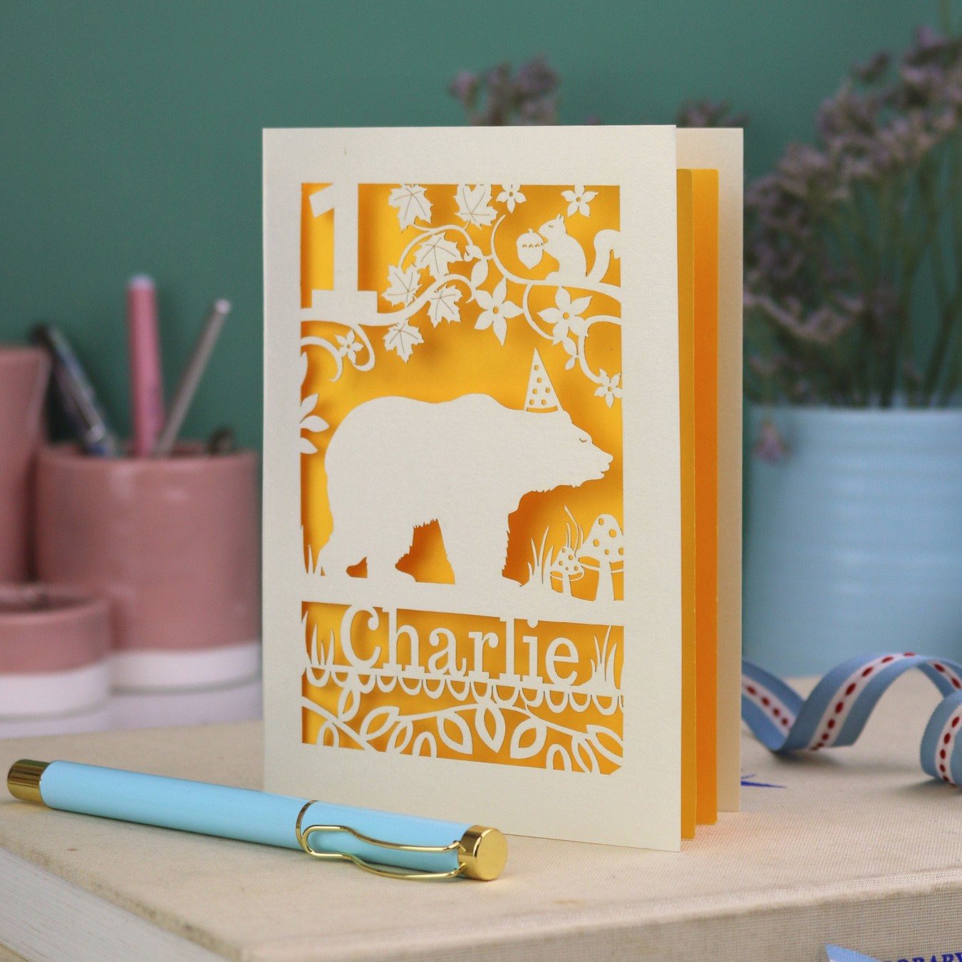 Personalised cut out 1st birthday card with bear design and 'Charlie' text on a table with stationery items.
