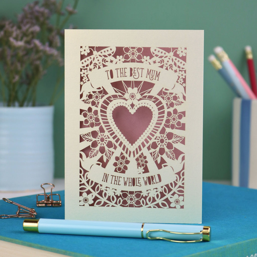 A laser cut card with intricate floral design with heart in the centre and the text "To the best mum in the whole world" cut out of two banners
