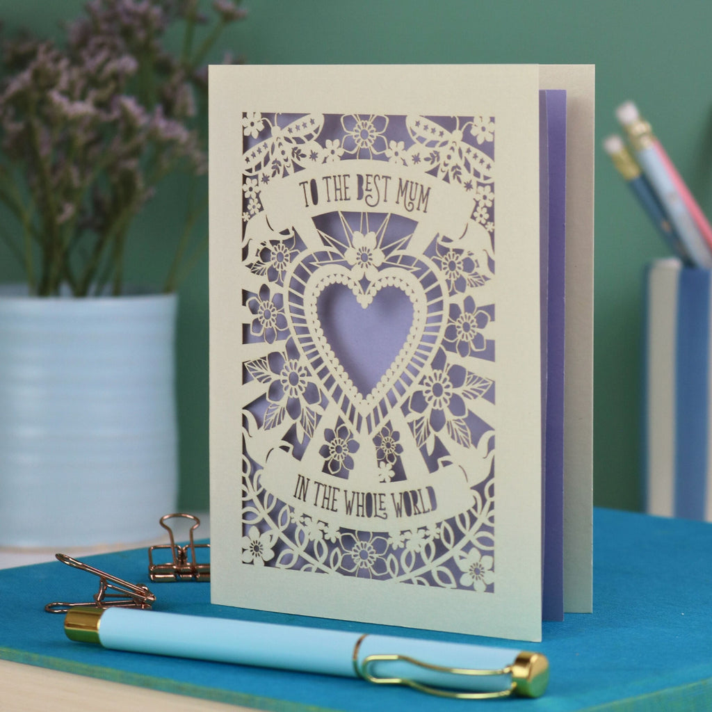 A laser cut card with intricate floral design with heart in the centre and the text "To the best mum in the whole world" cut out of two banners