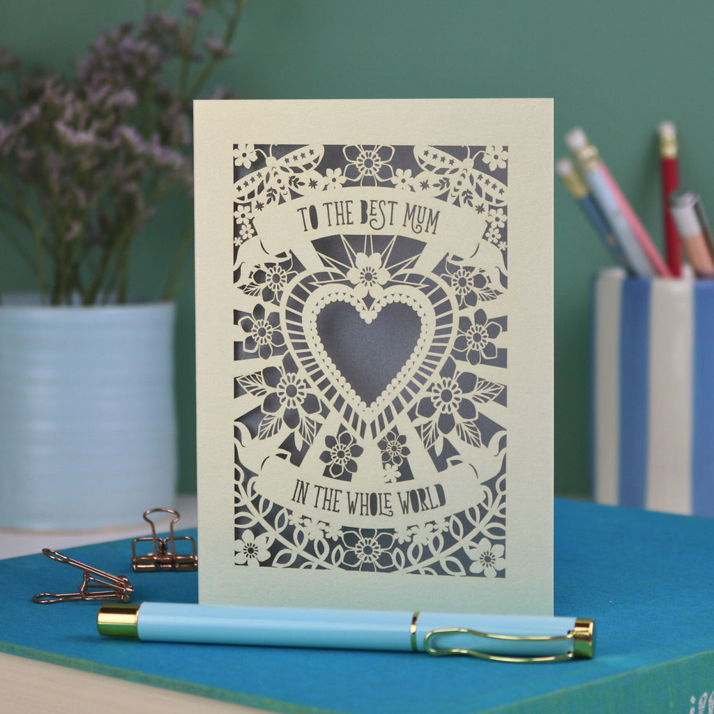 A laser cut card with intricate floral design with heart in the centre and the text "To the best mum in the whole world" cut out of two banners