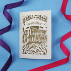 A personalised birthday card with the recipient's name laser cut from a banner shape and the words Happy Birthday in the middle, surrounded by floral designs. Birthday card is cream with gold leaf paper behind