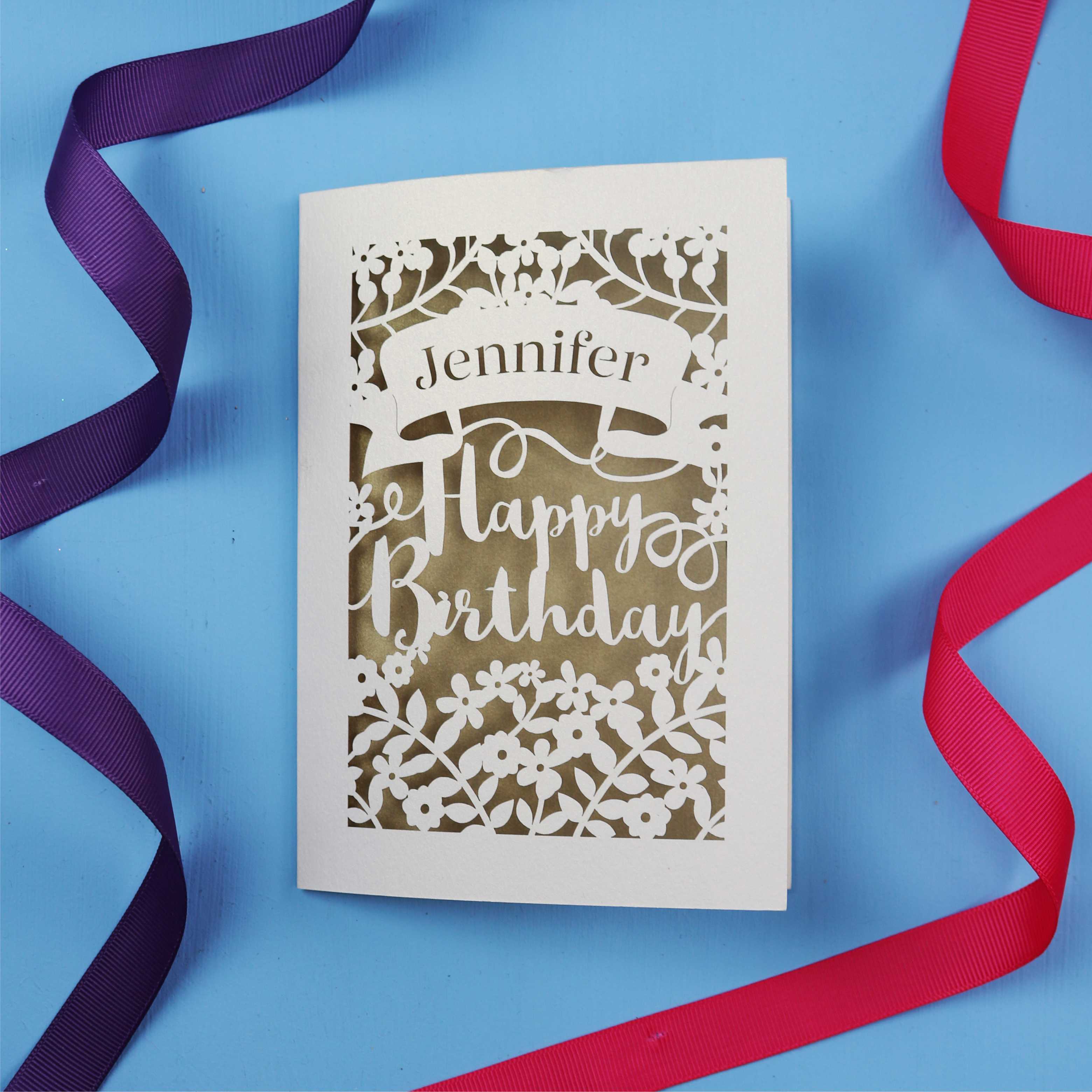A personalised birthday card with the recipient's name laser cut from a banner shape and the words Happy Birthday in the middle, surrounded by floral designs. Birthday card is cream with gold leaf paper behind