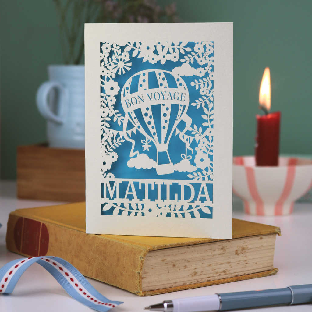 Card with 'Bon Voyage Matilda' design on a book with a candle and ribbon in the background