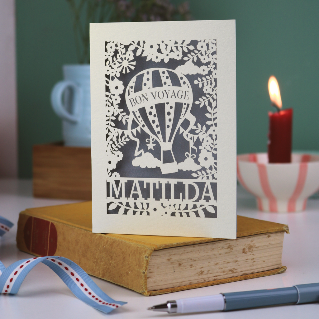 Laser-cut 'Bon Voyage Matilda' card on a book with a candle and ribbon in the background