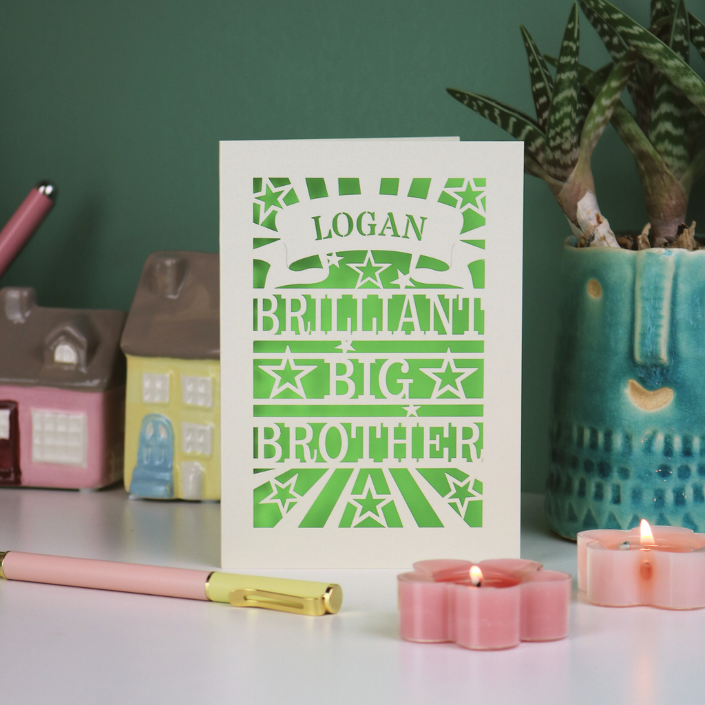 Card with 'LOGAN BRILLIANT BIG BROTHER' text on a table with candles and small houses.