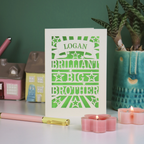 Card with 'LOGAN BRILLIANT BIG BROTHER' text on a table with candles and small houses.