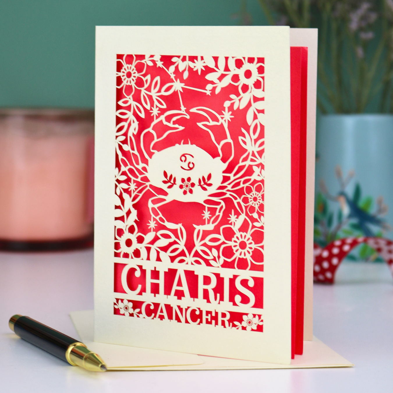 Decorative birthday card with cancer zodiac design and 'Charis', 'Cancer' text on a table with a pen and candle.