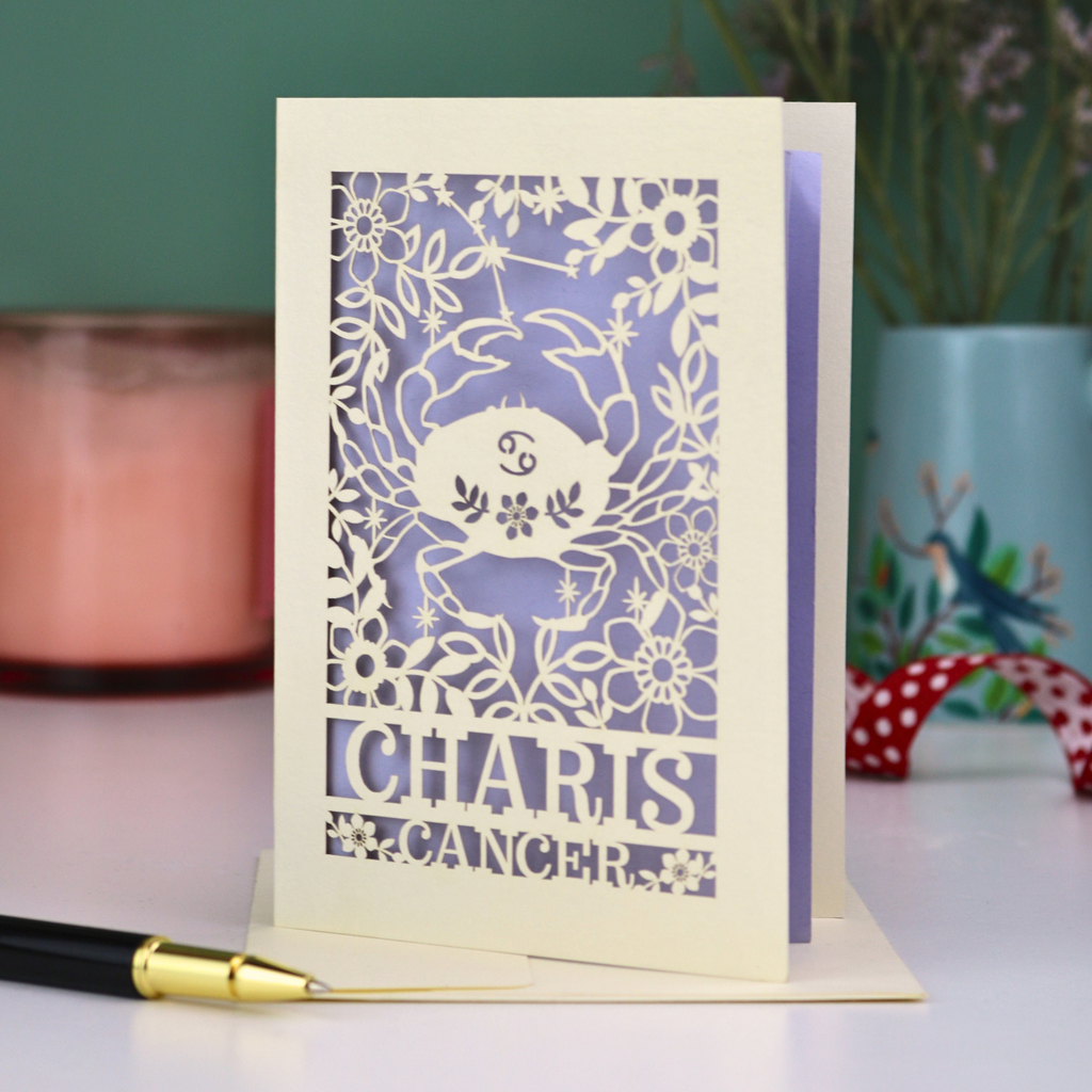 Decorative laser cut birthday card with cancer zodiac design and 'Charis', 'Cancer' text on a table with a pen and candle.