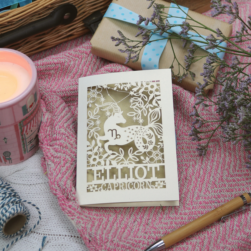 A laser cut Capricorn birthday card on pink background with candle, pen and gift. 