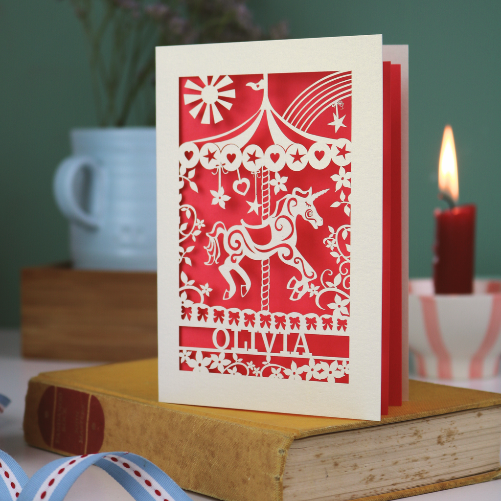 Decorative cut out birthday card with unicorn carousel design and personalised with a name, standing on a book with a blurred background