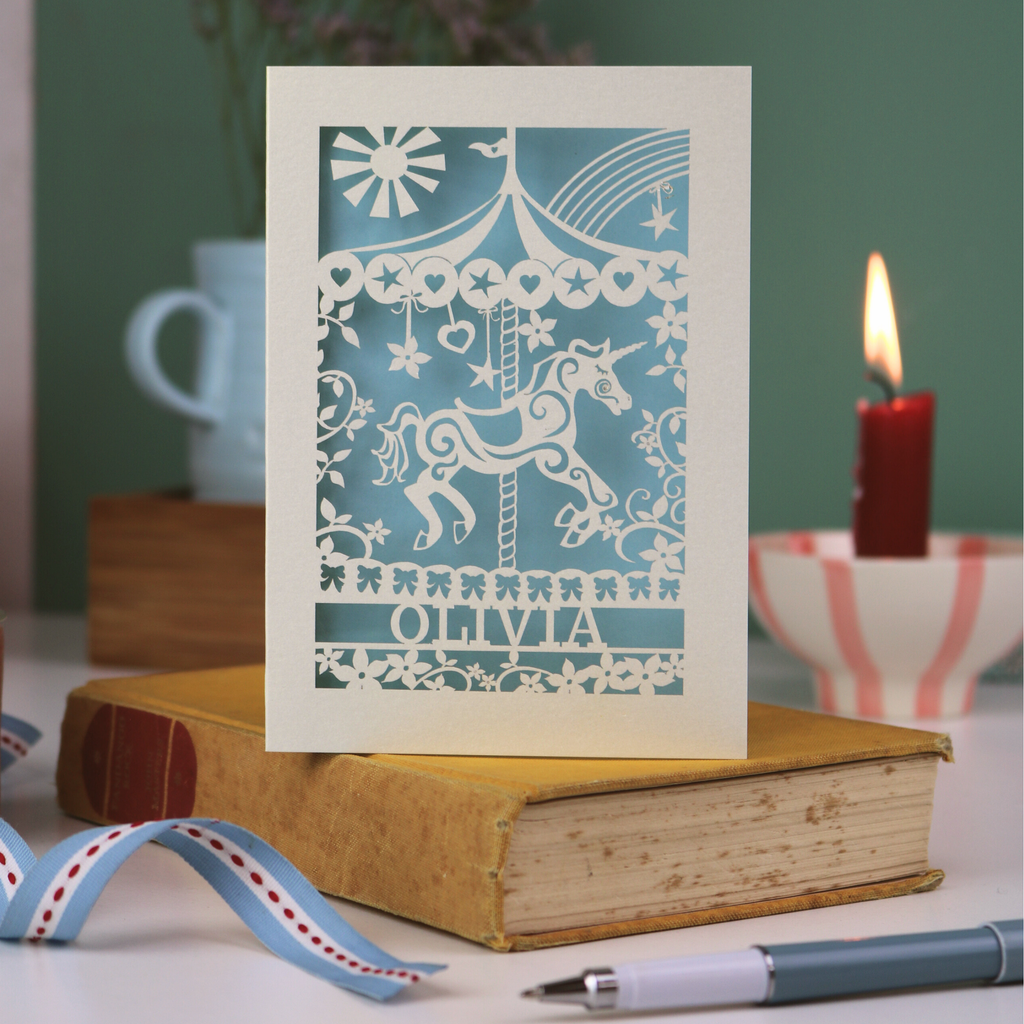 Decorative papercut birthday card with unicorn carousel design and personalised with a name, standing on a book with a blurred background