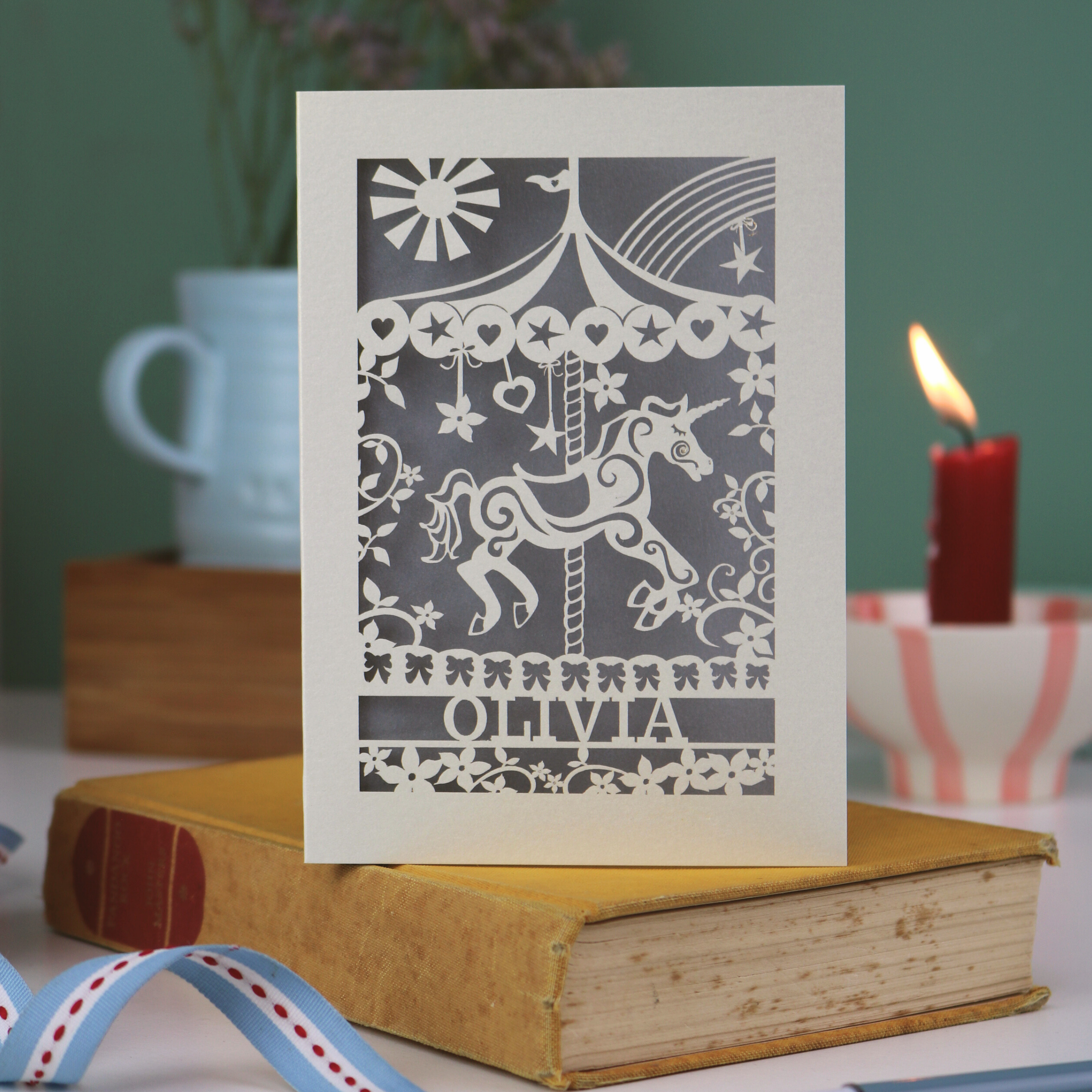 Decorative keepsake birthday card with unicorn carousel design and personalised with a name, standing on a book with a blurred background