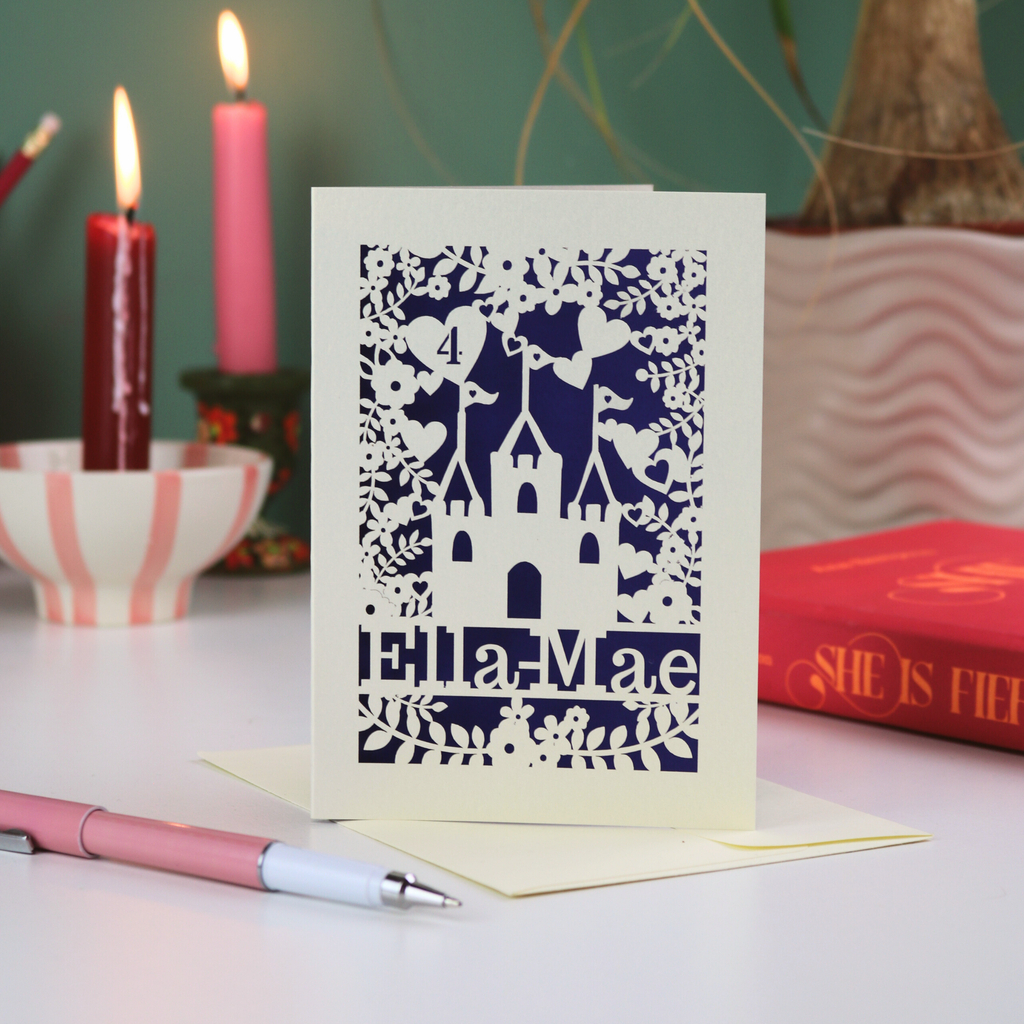 Decorative castle birthday  card with cut-out design and 'Ella Mae' text, placed on a white surface with candles and books in the background.