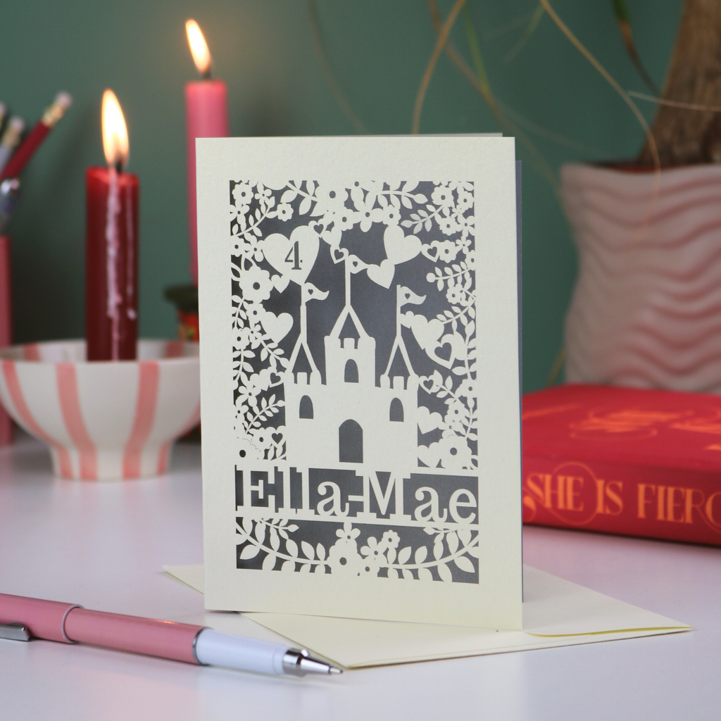 Decorative paper cut birthday card with cut-out floral and castle design and text, placed on a surface with candles and books in the background.