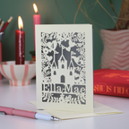 Decorative paper cut birthday card with cut-out floral and castle design and text, placed on a surface with candles and books in the background.
