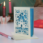 Laser-cut birthday card with castle design and 'Ella-Mae' text on a table with candles and books in the background.