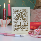 Laser-cut birthday card with castle design and 'Ella-Mae' text on a table with candles and books in the background.