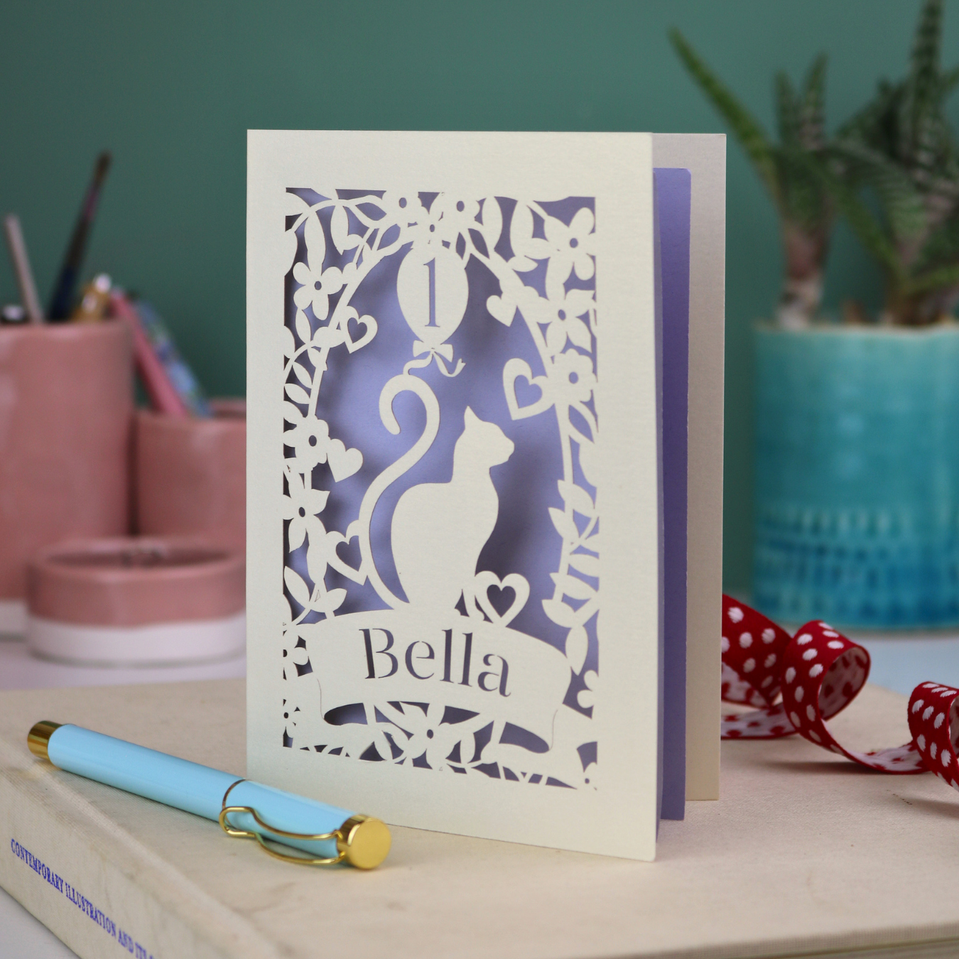 Laser cut 1st birthday card with a cat silhouette and 'Bella' text, placed on a surface with decorative items.