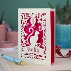 Paper cut first birthday card with cat design and 'Bella' text on a desk with a pen and decorative ribbon.