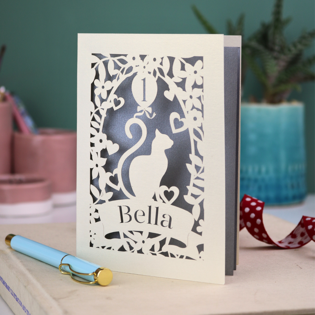 Laser-cut birthday card with a cat design and 'Bella' text on a table with a pen and decorative items.