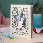 Laser-cut birthday card with a cat design and 'Bella' text on a table with a pen and decorative items.