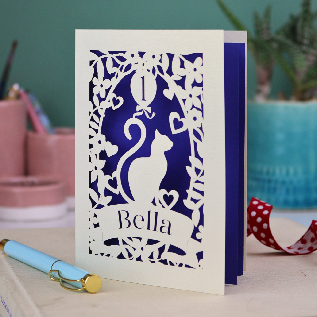 Personalised first birthday card with a laser cut cat silhouette and 'Bella' text, placed on a surface with decorative items.