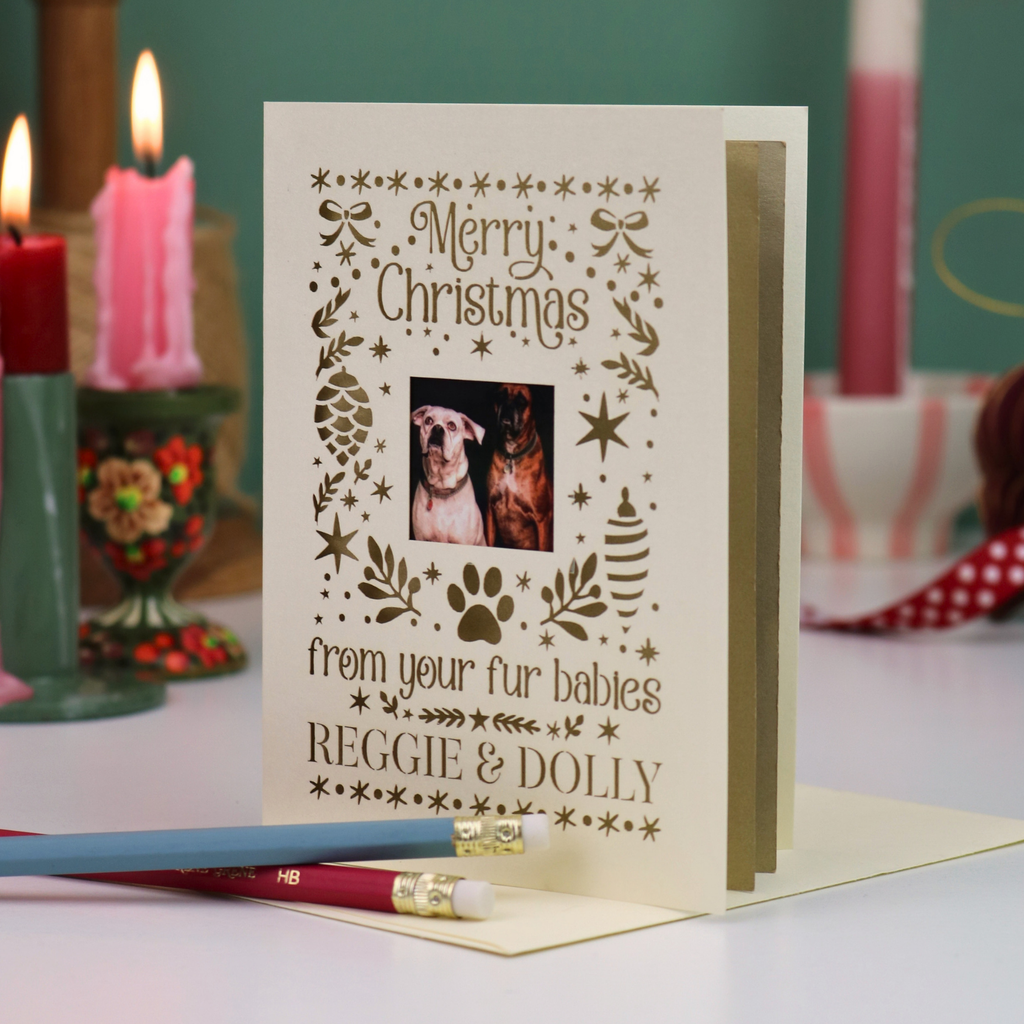 A Christmas greeting card with laser-cut holiday illustrations and a photo of two dogs, reading 'Merry Christmas from your fur babies REGGIE & DOLLY'. The card is displayed on a tabletop with candles and pencils in the background.
