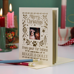 A Christmas greeting card with laser-cut holiday illustrations and a photo of two dogs, reading 'Merry Christmas from your fur babies REGGIE & DOLLY'. The card is displayed on a tabletop with candles and pencils in the background.