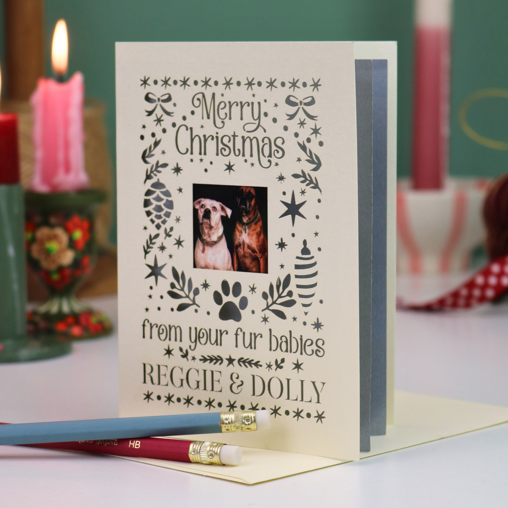 A personalized Christmas card with a photo of two dogs, reading 'Merry Christmas from your fur babies REGGIE & DOLLY', displayed on a table with a pencil in the foreground and holiday candles in the background.