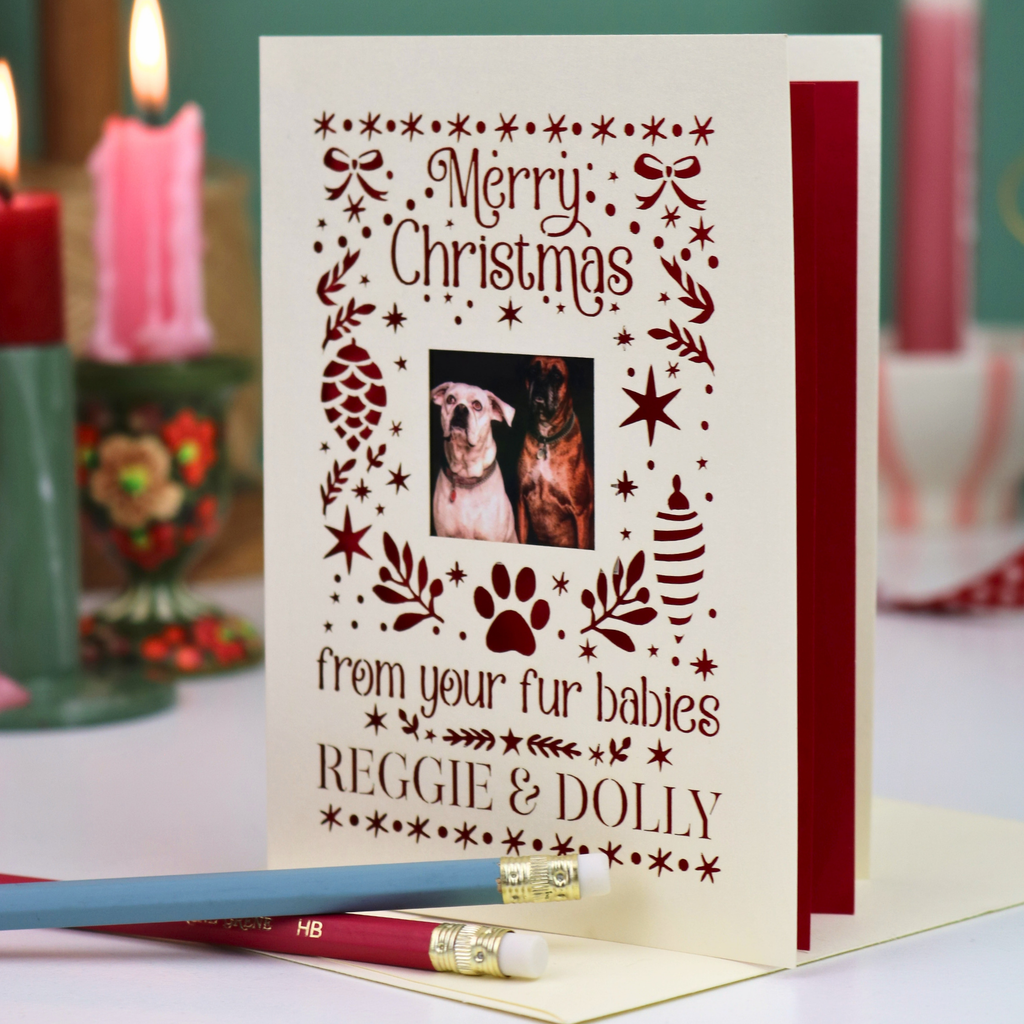 A personalized pet Christmas card with a photo of two dogs, printed with 'Merry Christmas from your fur babies REGGIE & DOLLY' in dark red. The card is shown standing open on a table with a pencil resting in front, with blurred holiday candles in the background.