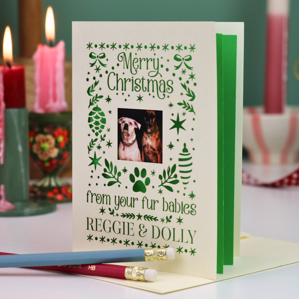 A personalized Christmas greeting card with festive border illustrations and a central photo of two dogs. The card is displayed open on a table with a pencil resting in front, and blurred candles are in the background.