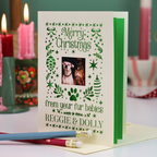 A personalized Christmas greeting card with festive border illustrations and a central photo of two dogs. The card is displayed open on a table with a pencil resting in front, and blurred candles are in the background.