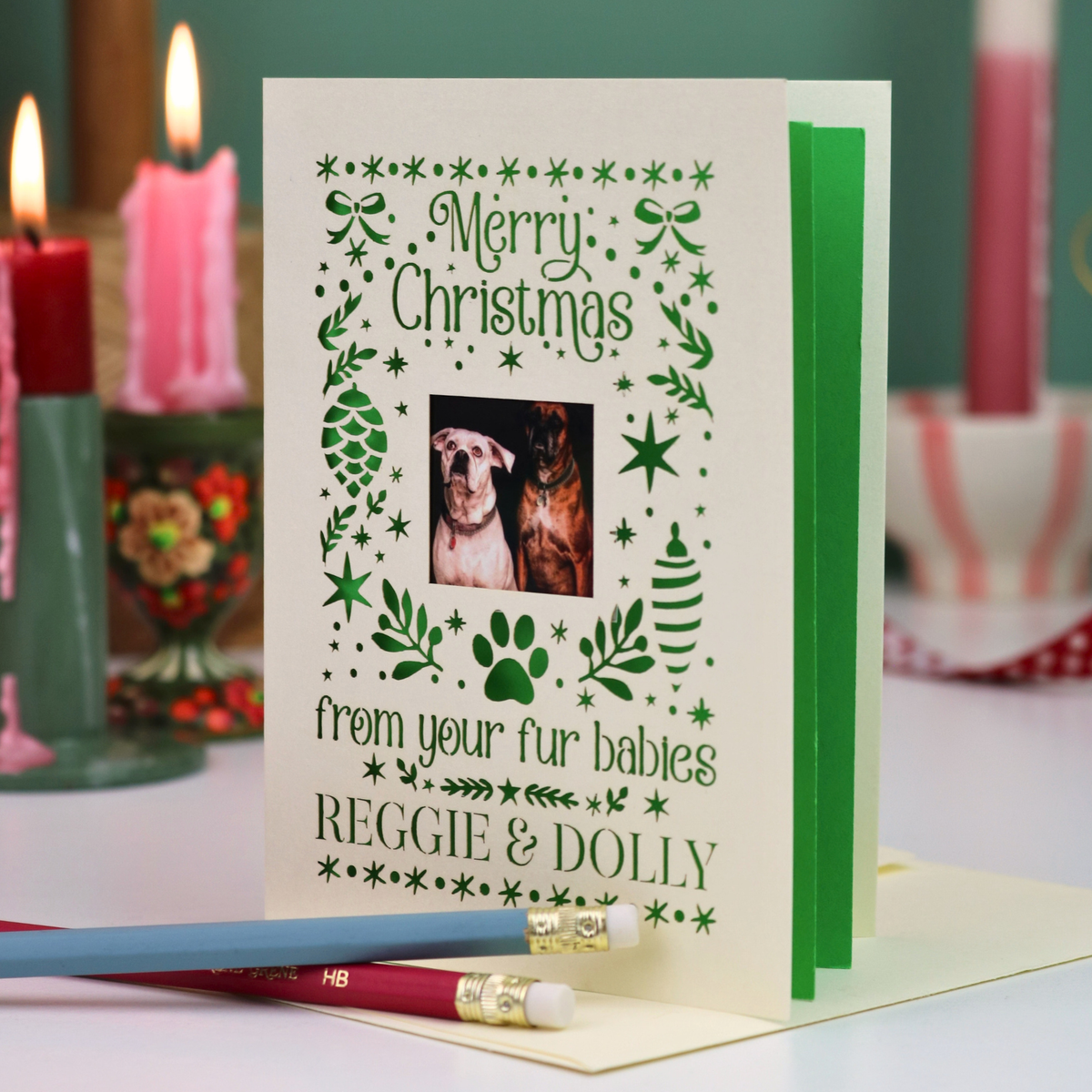 A personalized Christmas greeting card with festive border illustrations and a central photo of two dogs. The card is displayed open on a table with a pencil resting in front, and blurred candles are in the background.