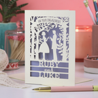 A laser cut engagement card featuring silhouettes of a couple making a heart-shape with their hands, with the text "on your engagement" and personalised with their names. Card sits on a white surface with stationery items, in front of a pink background. 