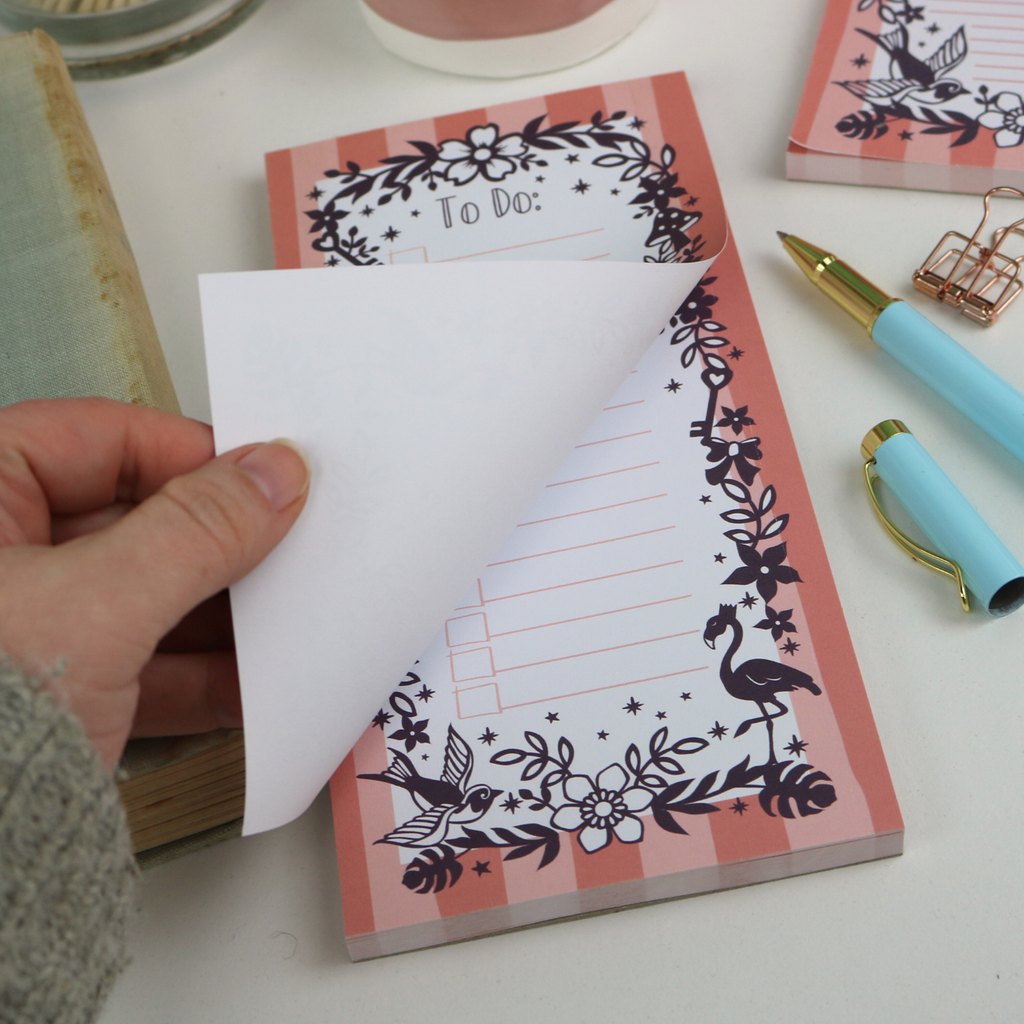 A close up detail of a notepad with pink striped border and purple floral illustrations, with "To Do:" at the top and lines with check boxes