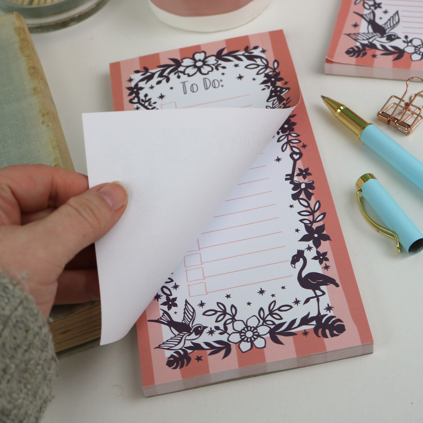 A close up detail of a notepad with pink striped border and purple floral illustrations, with "To Do:" at the top and lines with check boxes