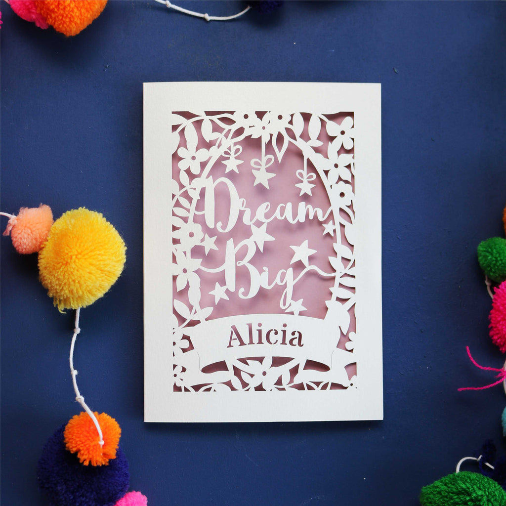 A personalised laser cut card with the words 'Dream Big' and a name 'Alicia' written on it, displayed on a dark background surrounded by colourful pompons.