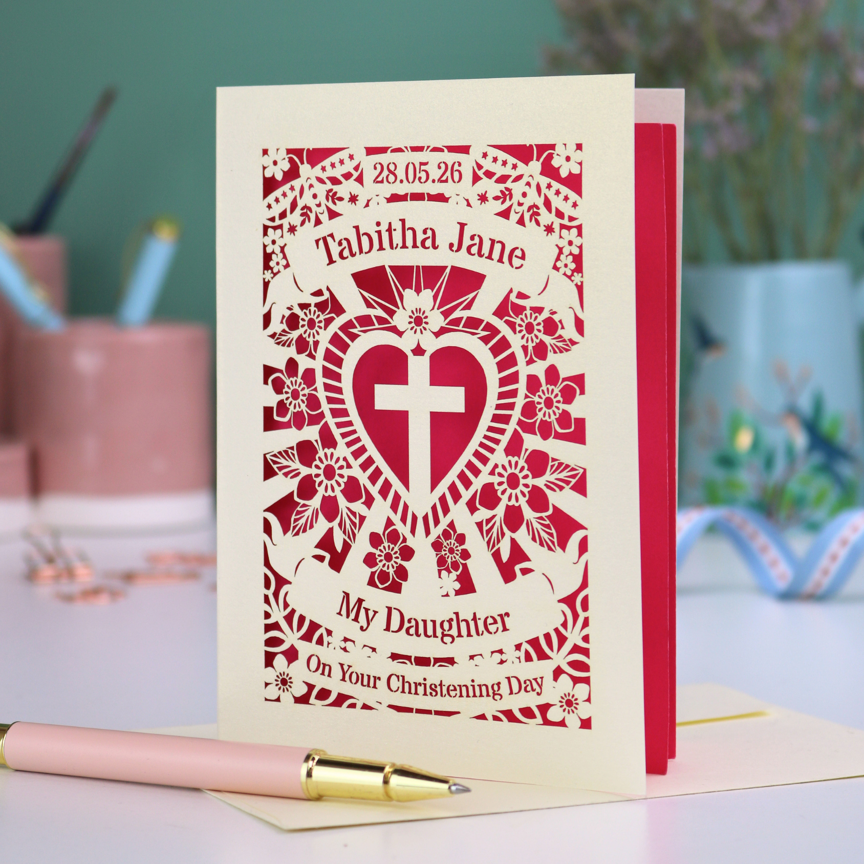 Personalized christening card with heart and cross laser cut design and text: "28.05.26, Tabitha Jane, My Daughter, On Your Christening day" on a desk with stationery items.