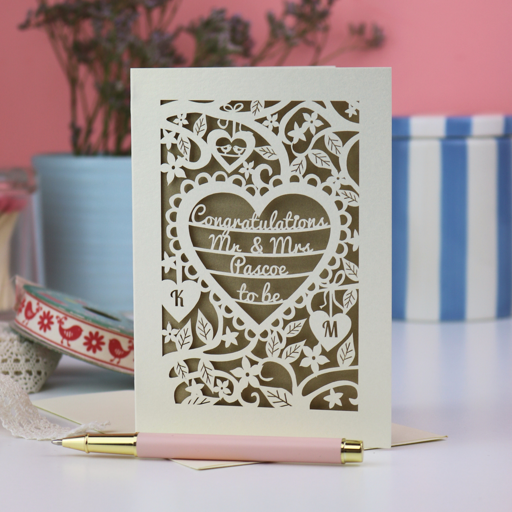 A laser cut engagement card with floral heart design, sitting on a white desk with ribbons, pen and pots on a pink background
