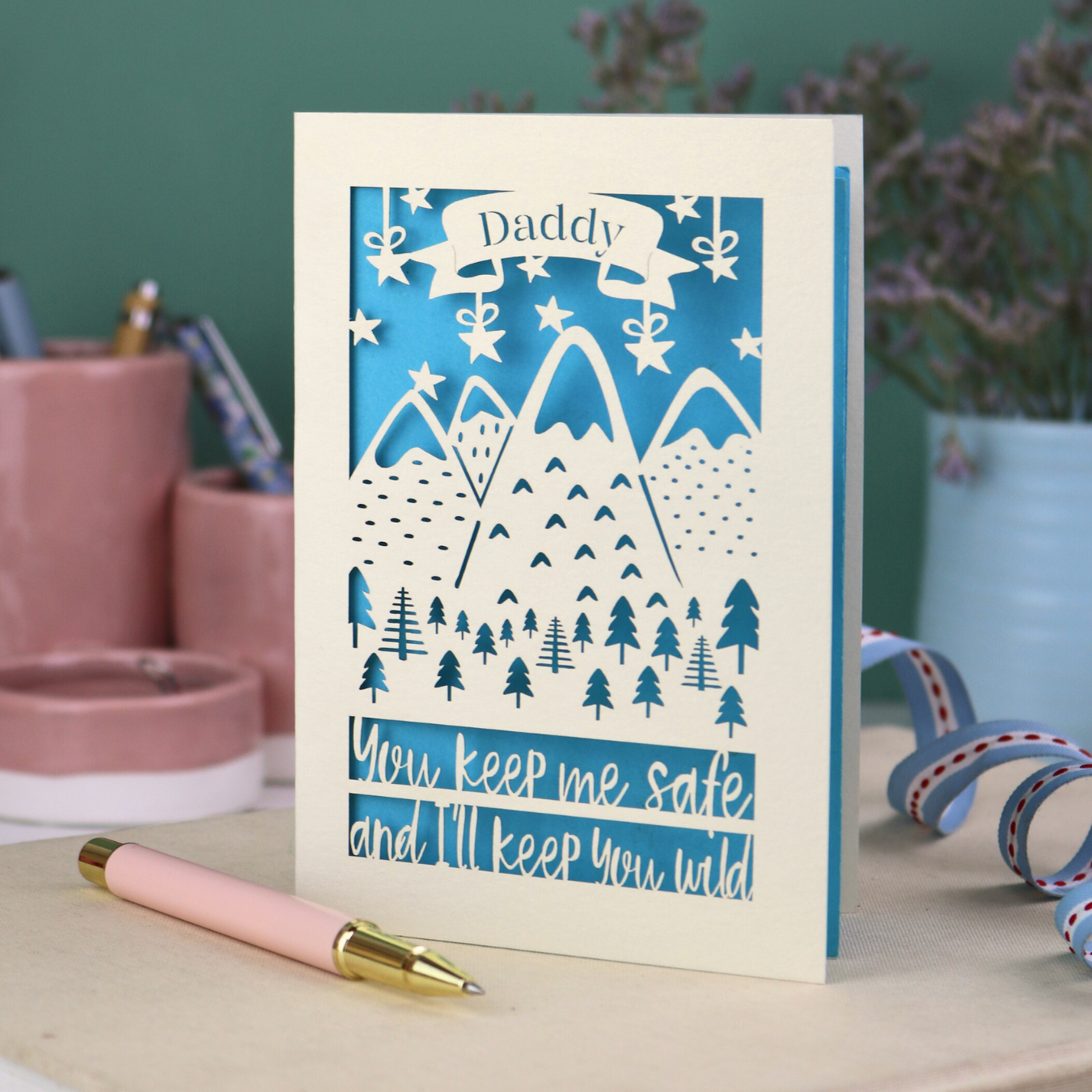 A cream laser cut personalised Father's Day card with mountain design and the text "Daddy" in a banner at the top, and "You keep me safe and I'll keep you wild" underneath
