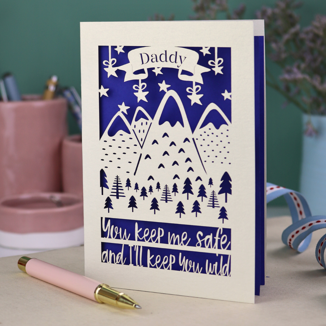 A cream laser cut personalised Father's Day card with mountain design and the text "Daddy" in a banner at the top, and "You keep me safe and I'll keep you wild" underneath