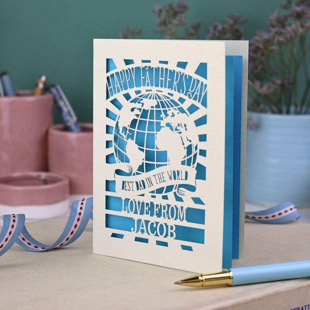 A laser cut Father's Day card with cream cut out card and a bright blue paper insert. Card has a globe design and the text "Happy Father's Day to the best dad in the world, love from" and is personalised with a name. 