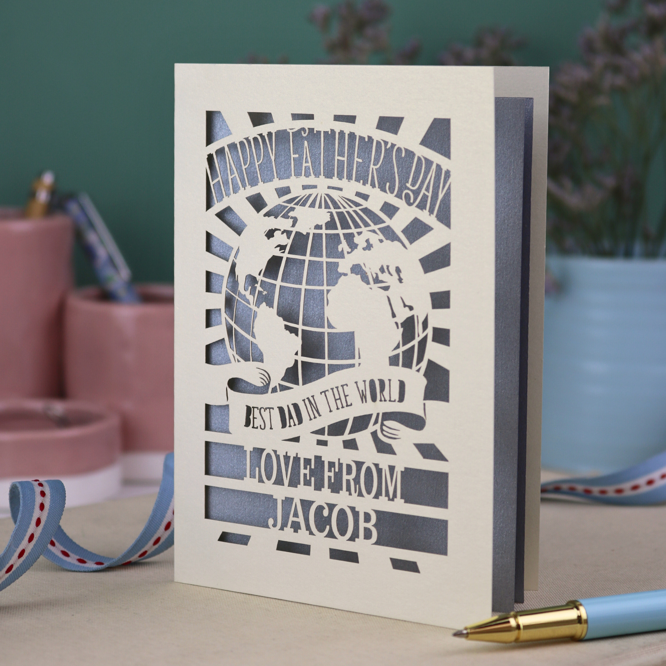 A laser cut Father's Day card with cream cut out card and a silver paper insert. Card has a globe design and the text "Happy Father's Day to the best dad in the world, love from" and is personalised with a name. 