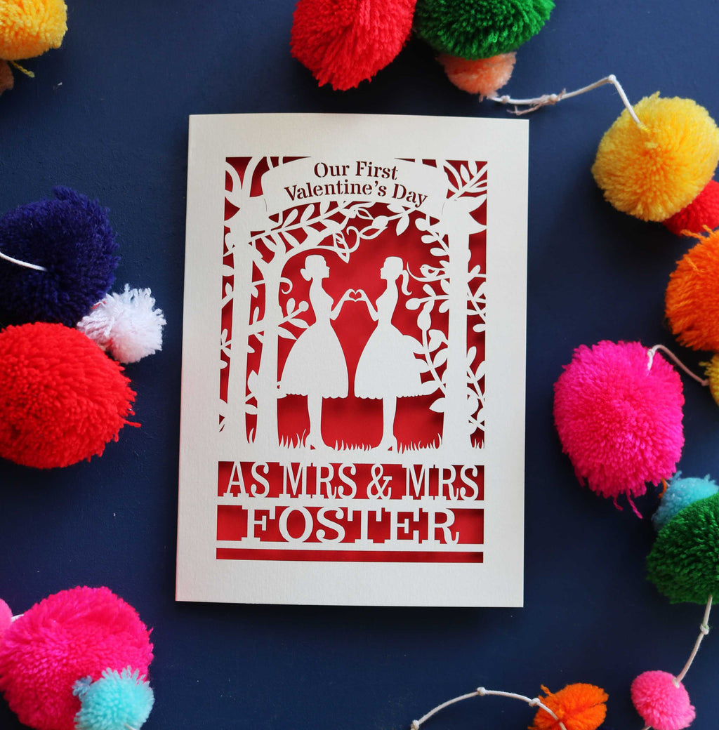 A personalised 1st Valentine's card with a laser cut design of two women making a heart shape, with a blank space for a personal message, and brightly coloured pom-pom decorations around it.