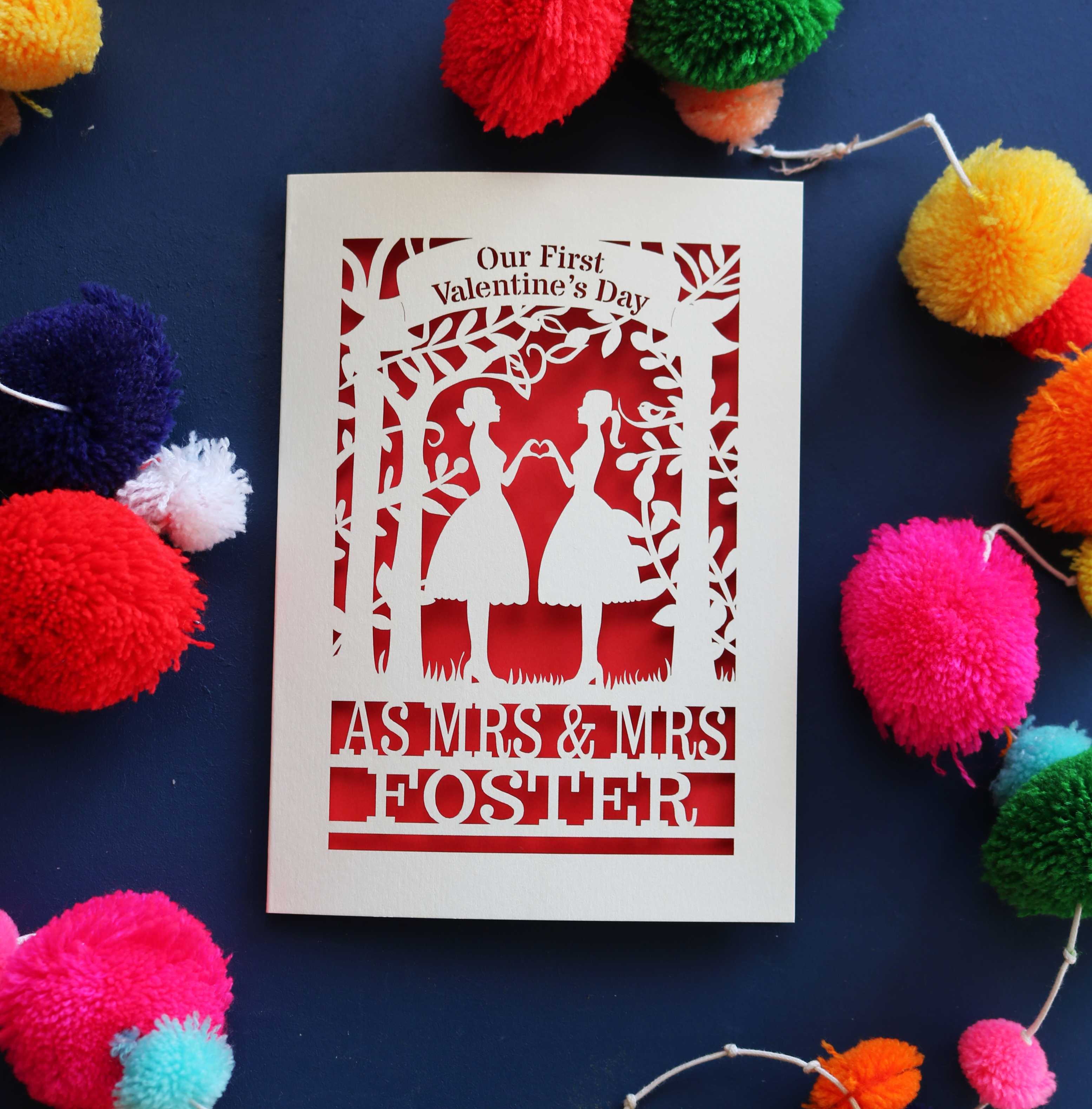 A personalised 1st Valentine's card with a laser cut design of two women making a heart shape, with a blank space for a personal message, and brightly coloured pom-pom decorations around it.
