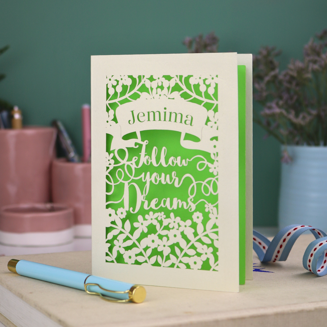 Decorative papercut graduation card with 'Jemima Follow Your Dreams' text on a desk with stationery items.