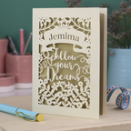 Decorative laser cut graduation card with 'Jemima Follow Your Dreams' text on a desk with stationery items.