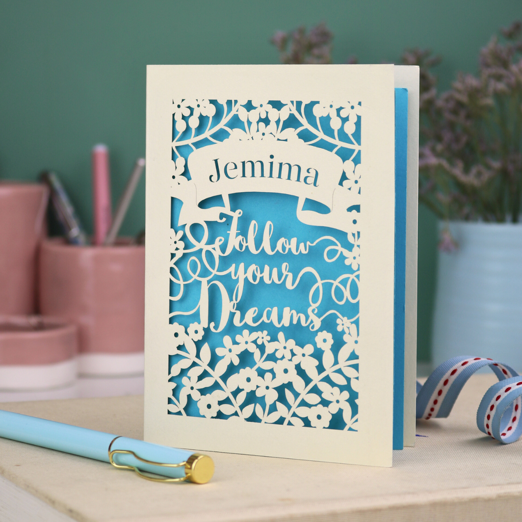Decorative personalised graduation card with 'Jemima Follow Your Dreams' text on a desk with stationery items.