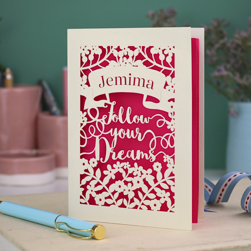 Decorative papercut graduation card with 'Jemima Follow Your Dreams' text on a desk with stationery items.