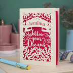 Decorative papercut graduation card with 'Jemima Follow Your Dreams' text on a desk with stationery items.